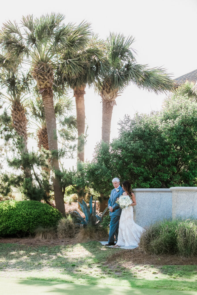 Father of the bride walks his daughter down the aisle underneath palm trees at the Bald Head Island Club.