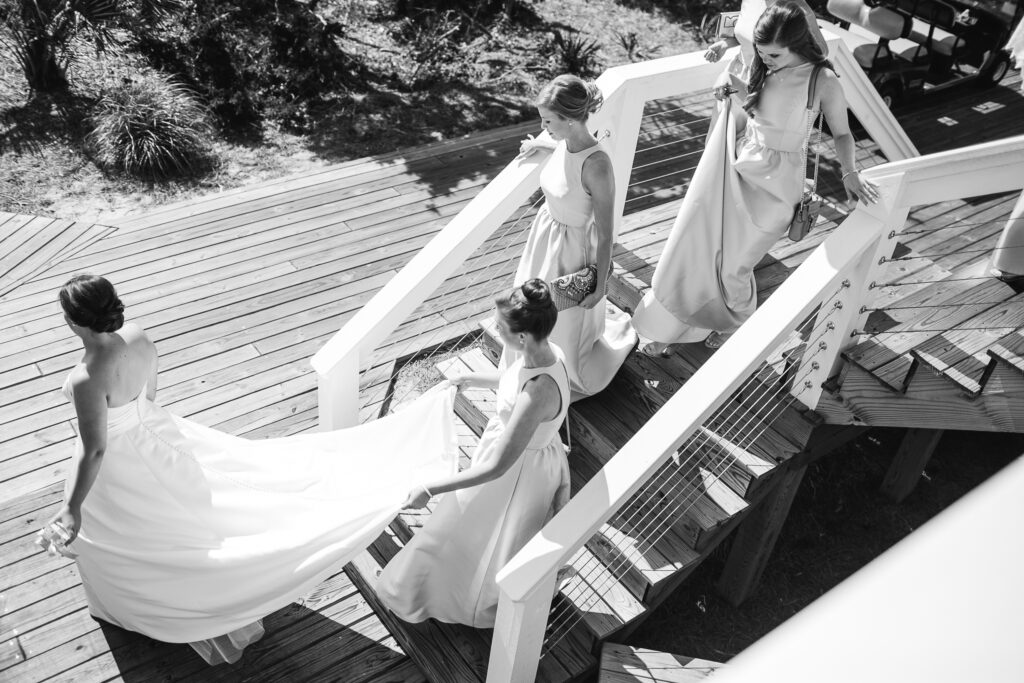 Bridesmaids help a bride walk down the steps by holding her dress as they walk towards her wedding ceremony on Bald Head Island.