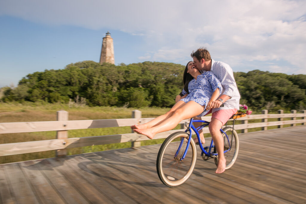 Engaged couple rides bike past Old Baldy Lighthouse on Bald Head Island during engagement session.